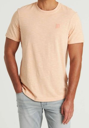 T-shirt à manches courtes de couleur pêche, en tissu doux, présentant une texture subtile et un petit écusson logo sur la poitrine gauche.