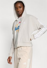 Ανοιχτό γκρι φούτερ με φερμουάρ και κουκούλα με κορδόνι, διακοσμημένο με λευκές ρίγες στα μανίκια και διακριτικές λογότυπες λεπτομέρειες, συνδυασμένο με μπεζ παντελόνια σε απόχρωση που ταιριάζει.