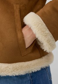 Bruine suède jas met een crèmekleurige nepbont voering aan de manchetten en zoom, met een zijzak zichtbaar aan de linkerkant.