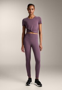Set sportiv de culoare mov, care include un top scurt cu maneci knotted și colanți cu talie înaltă. Materialul este neted și elastic, asortat cu teneși negri.