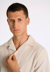 Camicia beige, con texture e colletto, dotata di una chiusura con un solo bottone. Il tessuto appare morbido e leggermente peloso.
