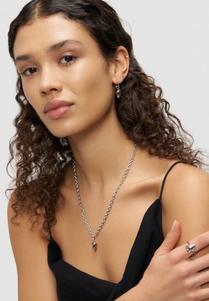 Junge Frau mit lockigem Haar trägt herzförmige silberne Ohrringe, dazu passenden Halskette und Ring, gekleidet in einem schwarzen Trägeroberteil.