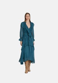 Türkises Maxikleid mit geschichteten Rüschen, langen, transparenten Ärmeln und einem Bindegürtel. Kombiniert mit beigen kniehohen Stiefeln für einen Kontrast.