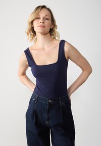 Bodysuit fără mâneci, de culoare albastru marin, cu decolteu pătrat, realizat dintr-un material neted. Asortat cu pantaloni din denim închis, cu talie înaltă.
