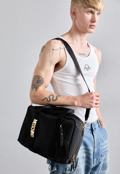 Jeune homme avec des tatouages portant un débardeur blanc et un jean bleu, portant un sac à bandoulière noir Moschino avec des accents dorés.