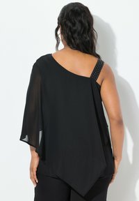Asymmetrisk sort bluse med løs pasform, gennemsigtigt stof og en besat rem. Har en flydende ryg og skrå søm.