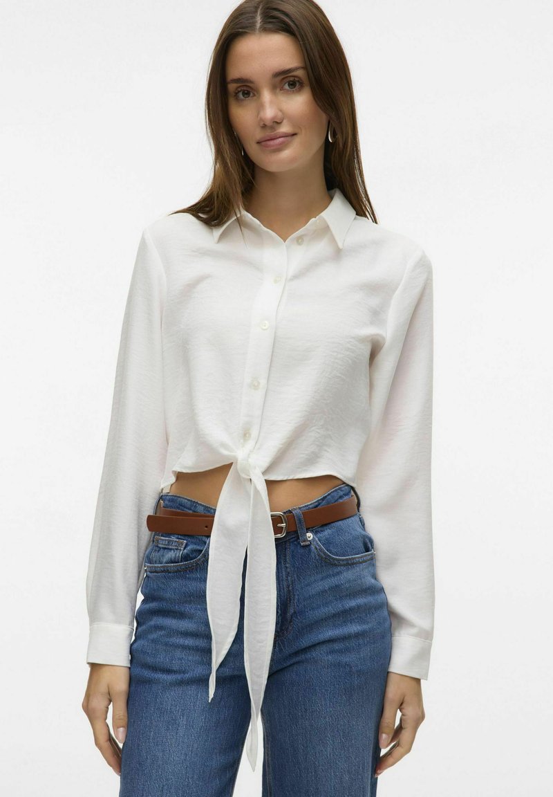 Camicia bianca con bottoni e un nodo in vita, caratterizzata da maniche lunghe e un tessuto testurizzato, abbinata a jeans blu a vita alta e una cintura marrone.