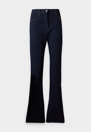 Persona in piedi che indossa un maglione bianco con ampie righe blu navy, pantaloni neri a vita alta svasati e scarpe rosso scuro, mani nelle tasche.