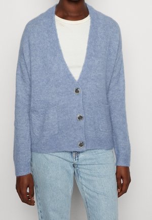Personne portant un cardigan bleu clair boutonné avec des poches frontales par-dessus une chemise blanche et un jean bleu clair, devant un fond uni.