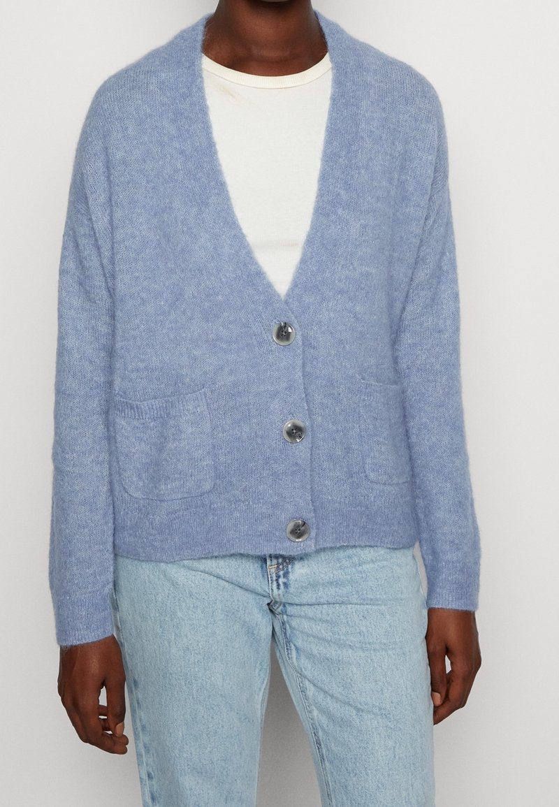 Personne portant un cardigan bleu clair boutonné avec des poches frontales par-dessus une chemise blanche et un jean bleu clair, devant un fond uni.