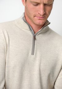 Helle beige Strickjacke mit einem halben Reißverschluss-Kragen, ausgestattet mit einer gerippten Textur und einem kontrastierenden grauen Reißverschluss sowie Detail im Inneren des Kragens.