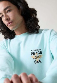 Sudadera azul claro hecha de tela suave que presenta un diseño gráfico central con el texto "ALTONADOCK PAZ AMOR MAR" y acentos alados.