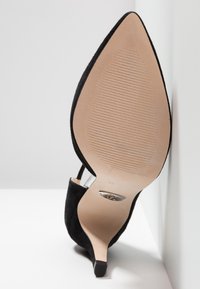 Chaussure à talon haut en daim noir à bout pointu vue par en dessous, mettant en avant la semelle beige avec des lignes de grip texturées et une petite plaque métallique.