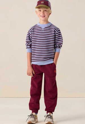 Gestreepte paarse en blauwe sweatshirt met ronde hals, gecombineerd met bordeauxrode joggingsbroek met een ronde patch. Grijze en bruine sneakers maken de look compleet.