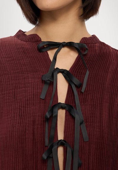 Burgundijas krāsas tops ar ribota tekstūra, ar V formas izgriezumu un melnas satīna lentes mezgliem pie apkakles. Audums izskatās mīksts un nedaudz sakļauts.