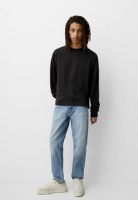 Mladić s dugom kovrčavom kosom nosi crnu majicu dugih rukava, svijetloplave traperice i bijele tenisice, stojeći uz bijeli zid.