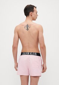 Rosa Badeshorts mit einem schwarzen elastischen Bund, der mit dem "CALVIN KLEIN" Branding versehen ist. Die Shorts haben eine Gesäßtasche und eine glatte Textur.