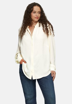 Chemise à manches longues, boutonnée, de couleur crème, en tissu texturé, avec une coupe décontractée, détail de poche et poignets retroussés, associée à un jean en denim foncé.