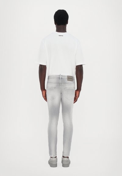 T-shirt en coton blanc avec un logo dans le dos, associé à un jean en denim gris au look déchiré et à un patch logo à l'arrière, porté avec des chaussures grises.