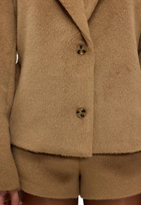 Blazer en mélange de laine couleur sable avec un col à revers cranté, doté de deux boutons. Associé à un short assorti avec une finition douce et texturée.