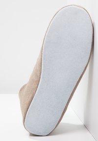Beige suède tøfler med en lys blå gummisåle, som har en myk tekstur og rund form, vist fra en sidevinkel mot en hvit bakgrunn.