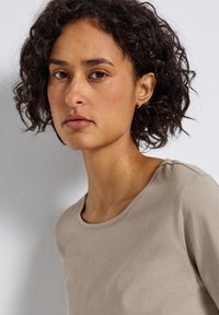 Donna con capelli ricci scuri che indossa una camicia beige, guardando leggermente di traverso su uno sfondo chiaro e semplice.