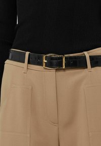 Suéter negro acanalado combinado con pantalones beige con bolsillos laterales, sujetos con un cinturón negro brillante de estampado cocodrilo y hebilla dorada.