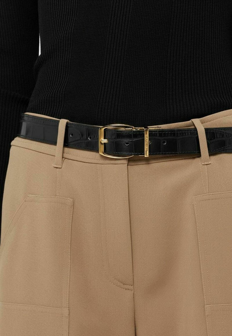Suéter negro acanalado combinado con pantalones beige con bolsillos laterales, sujetos con un cinturón negro brillante de estampado cocodrilo y hebilla dorada.