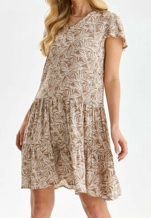 Beige kortærmet kjole med lagdelt design og hvidt bladmønster, båret af en kvinde med blondt hår, stående foran en ensfarvet hvid baggrund.