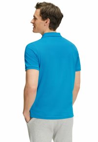 Polo blu con colletto corto, texture liscia e maniche corte. Indossato con pantaloni della tuta grigi, che mostrano un fit e un design rilassati.