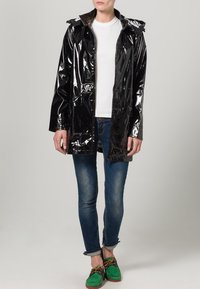 Musta kiiltävä sadetakki hupulla, vetoketjulla, kahdella etutaskulla, valkoisen paidan päällä, yhdistetty sinisiin farkkuihin ja vihreisiin kenkiin.