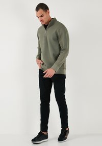 Hanorac cu fermoar de culoare verde măsliniu, cu mâneci lungi, manșete ribbate și guler înalt, asortat cu pantaloni negri slim și adidași negri.