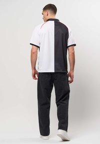 Homme debout, de dos, portant un polo manches courtes moitié noir, moitié blanc, un jean noir et des baskets blanches sur un fond uni.