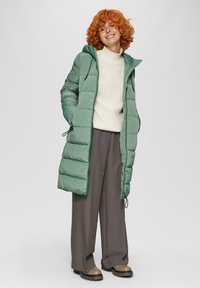Manteau rembourré long vert avec capuche, présentant un design matelassé et un cordon de serrage. Porté sur un pull crème et un pantalon gris, associé à des chaussures marron.