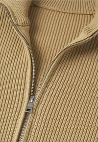 Tissu en maille côtelée beige avec une fermeture éclair en métal argenté partiellement ouverte et un col plié sur un vêtement.