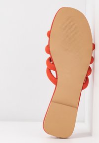 Scarpa in suede rosso con suola piatta, caratterizzata da una superficie liscia e texture e anelli decorativi lungo il design superiore. La suola è di colore beige e piatta.
