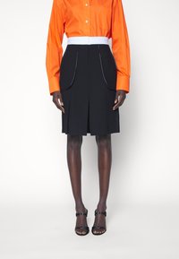 Oranssi napitettava paita, jossa on pitkät hihat, yhdistetty mustaan hameeseen, jossa on sivutaskut ja valkoiset ompeluyksityiskohdat. Mustat korkokenkien sandaalit.