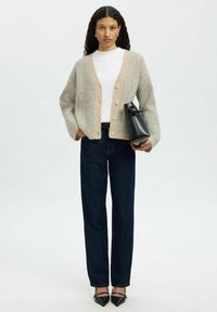 Vaalean beige ribbed-neulecardigan, jossa on V-pääntie ja napit, yhdistetty valkoiseen paitaan, tummansiniset farkut ja teräväkärkiset mustat korkokengät; pitäen mustaa laukkua.