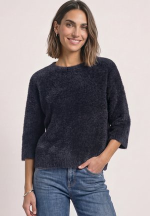 Femme souriante aux cheveux mi-longs portant un pull sombre et duveteux et un jean bleu, une main dans la poche.