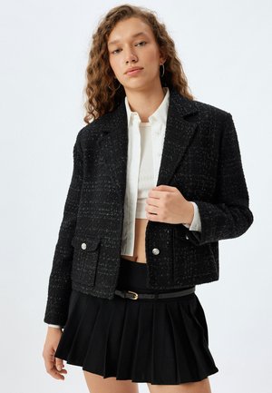 Blazer en tweed noir avec un design court, deux poches avant et des détails en boutons argentés. Porté sur une chemise blanche et associé à une jupe plissée noire.