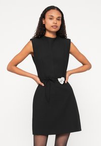 Schwarzes ärmelloses Kleid mit tailliertem Oberteil, betontem Bund und einer dekorativen silbernen Brosche. Glattes Material und eine kurze, A-Linie Silhouette.