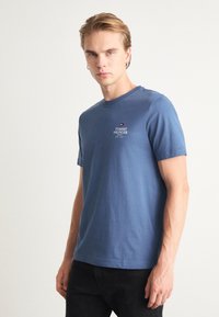 T-shirt in cotone blu con maniche corte, colletto a girocollo e un piccolo logo ricamato sul petto. Indossato con jeans neri.