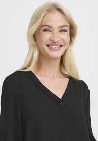 Blond kvinde smilende, iført en sort V-hals top med en enkelt knap, mod en ensfarvet lys baggrund.