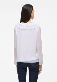 Hvit langermet bluse med volangkrage og glatt tekstur, kombinert med mørkeblå tettsittende jeans.