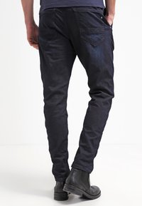 Jeans aderenti blu navy scuro con una leggera lucentezza, dotati di tasche posteriori e una texture liscia. Abbinati a stivaletti neri alla caviglia.