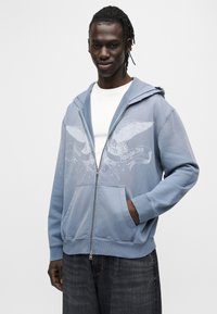 Φούτερ με φερμουάρ σε ανοιχτό γαλάζιο χρώμα, με μπροστινή τσέπη, που διαθέτει γραφικό σχέδιο με φτερά και κείμενο. Μαλακή υφή, με ριμπ μανσέτες και τελείωμα.