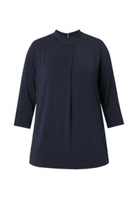 Navy sininen paita, jossa on korkea kaulus, kolmannen neljänneksen hihat, pehmeä kangas, etupuolella laskos yksityiskohtana ja pieni vetoketju takana.