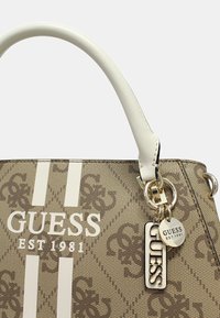 Bolso beige con rayas blancas y "GUESS EST 1981" impreso en letras negritas, con un llavero de logo plateado adjunto.