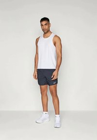 Valkoinen hihaton t-paita, tummansiniset shortsit, joissa on pieni valkoinen Nike-logo, ja valkoiset urheilukengät; malli seisoo yksinkertaisella taustalla.