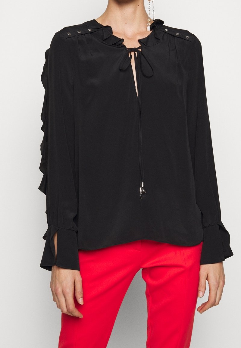 Blouse noire avec col froncé, devant noué et accents de boutons sur les épaules. Manches longues avec des poignets fendus. À porter avec un pantalon rouge ajusté.